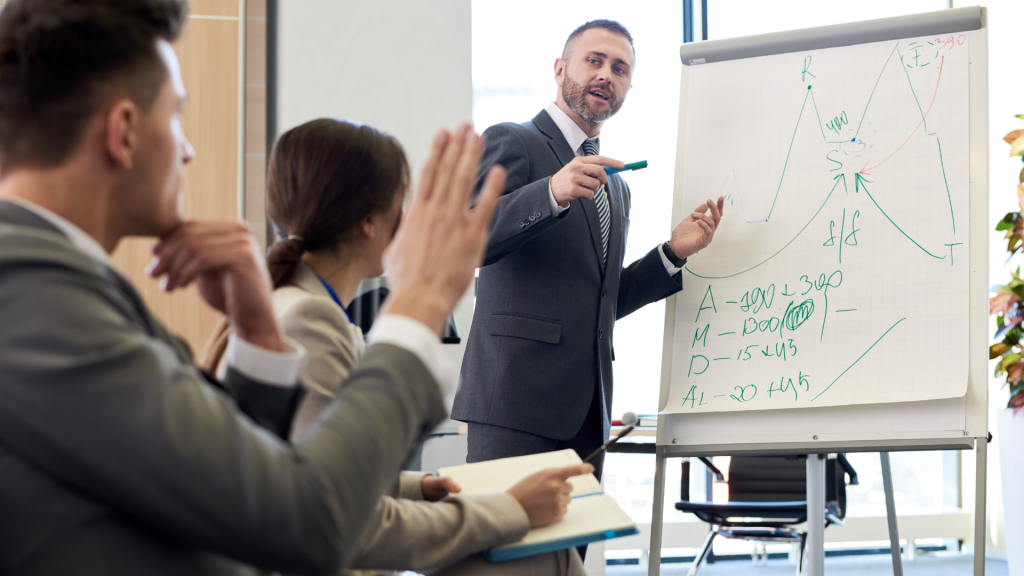Bearded telecom instructor standing by whiteboard explaining concepts to telecom executives while one raises his hand to ask a question
