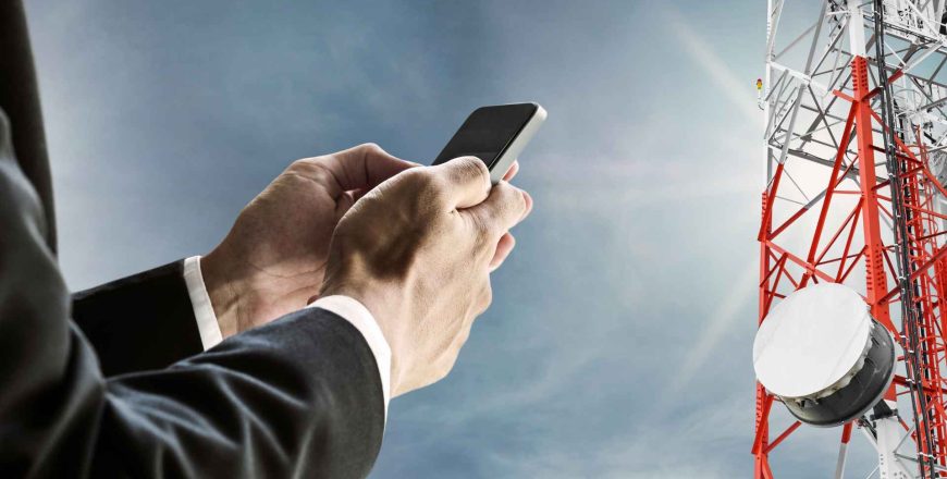 Businessman using mobile phone, with satellite dish telecom network on telecommunications tower against blue sky