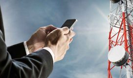 Businessman using mobile phone, with satellite dish telecom network on telecommunications tower against blue sky